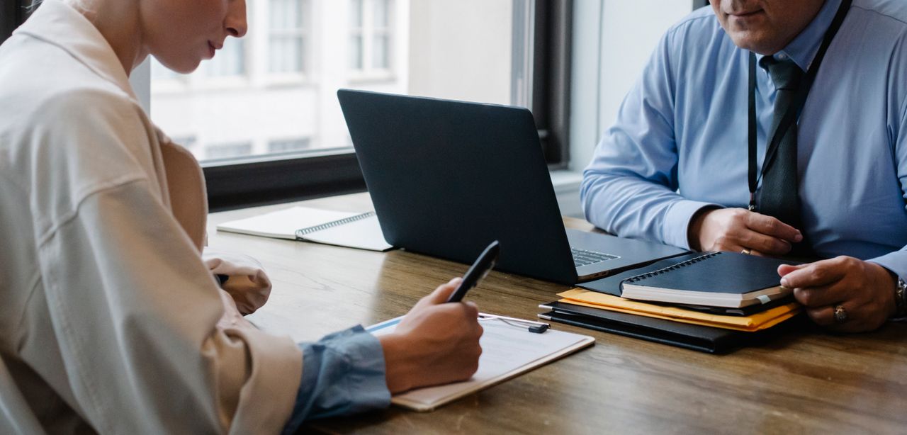 Crop faceless colleagues in formal clothes sitting at table with laptop and folder and signing documents while working together
